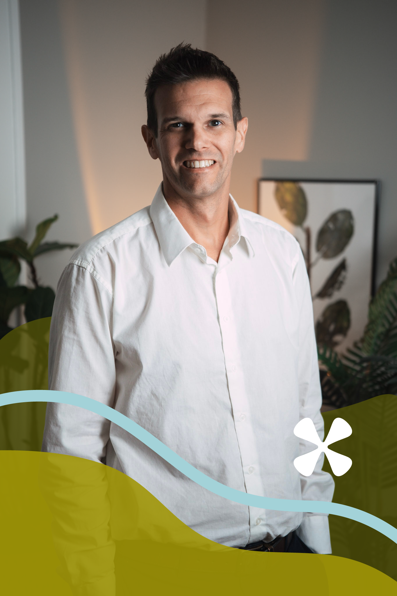 A man in a white shirt smiling in an indoor setting with plants and framed artwork in the background.