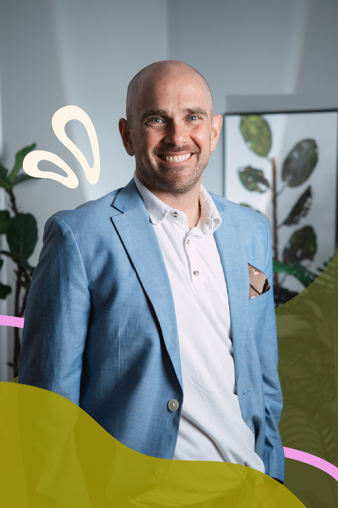 A smiling man with a shaved head and blue eyes, dressed in a light blue blazer and white shirt, standing indoors with plants and a picture frame visible in the background.
