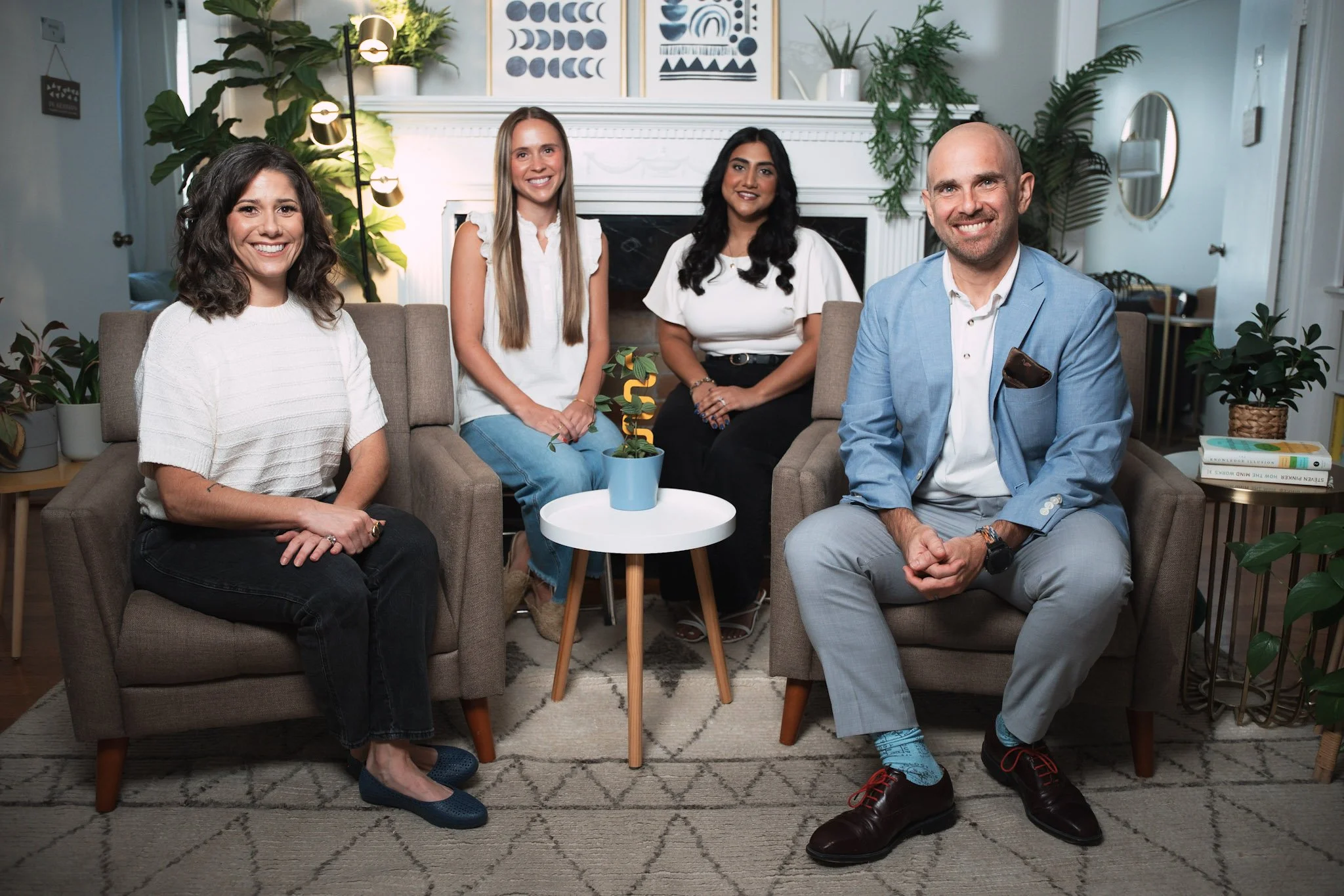 Five people sitting on a sofa in a cozy living room, smiling at the camera. The room has plants, framed artwork, and books, with a small white table holding a potted plant in front of them.