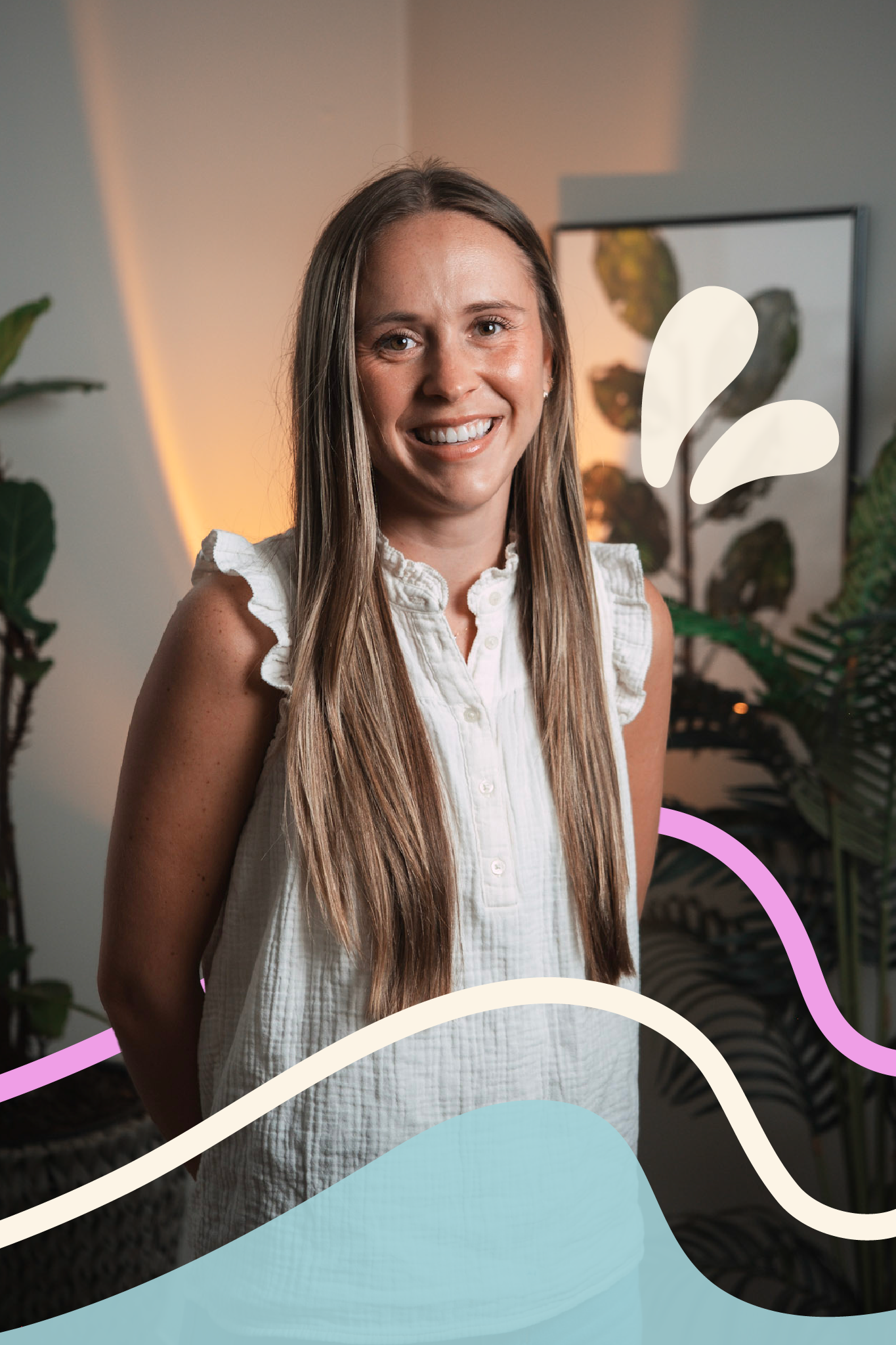 A woman with long brown hair is smiling and standing in front of a wall with framed plant artwork. She is wearing a sleeveless white blouse with ruffles.