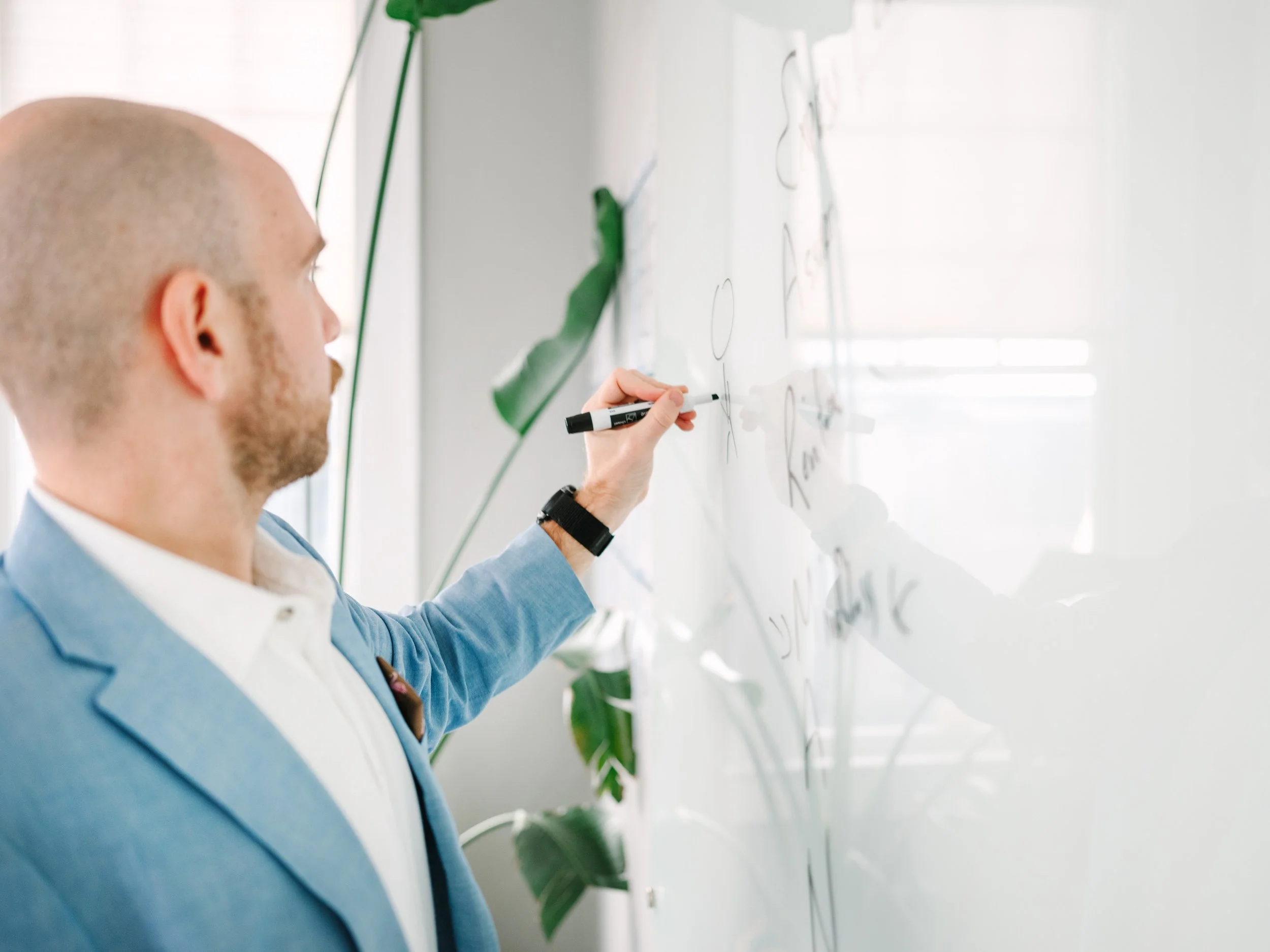 A man with a shaved head and a beard, dressed in a light blue blazer and white shirt, is writing on a whiteboard with a black marker in a classroom or office. There are green plants and a window with white blinds in the background.