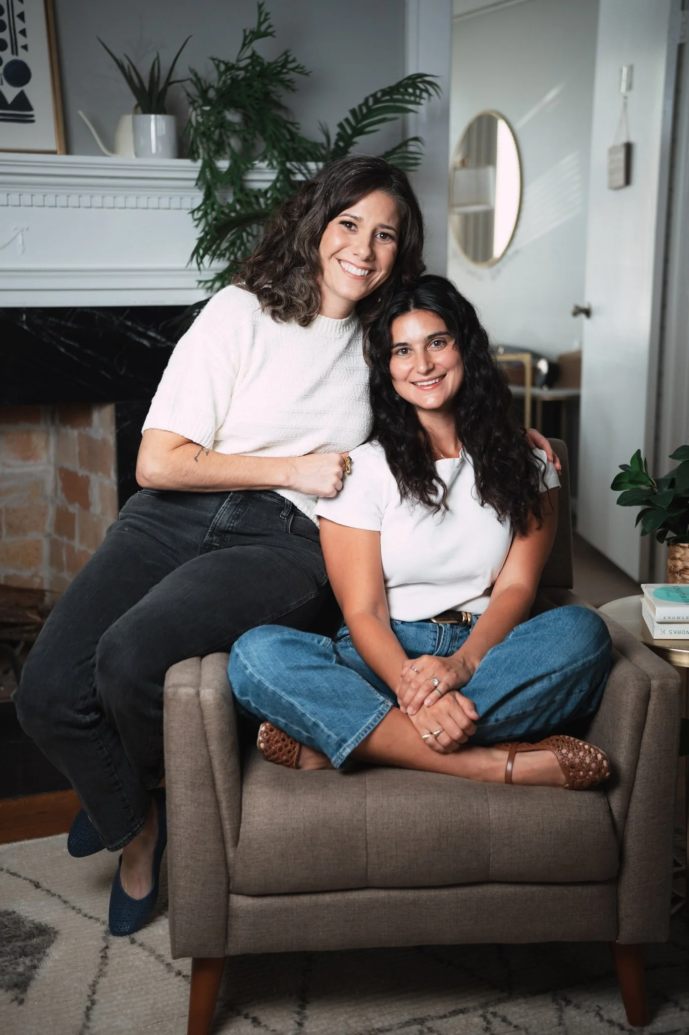 Two women smiling, sitting on a gray armchair in a cozy living room. One woman with curly dark hair is seated, wearing a white t-shirt and jeans, and the other woman with wavy brown hair is standing beside her, wearing a white sweater and black pants. There are houseplants and a fireplace behind them.