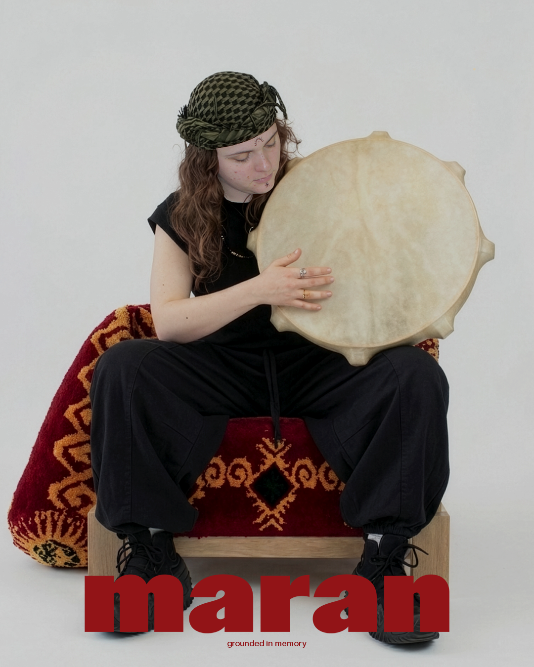 A young person with long hair, wearing a patterned headscarf, black clothing, and black shoes, sits on a decorative red and gold cushion, holding a large, round drum with a beige surface against their chest. The background is plain white. The word 'maran' in large red letters and the phrase 'grounded in memory' are overlaid at the bottom of the image.
