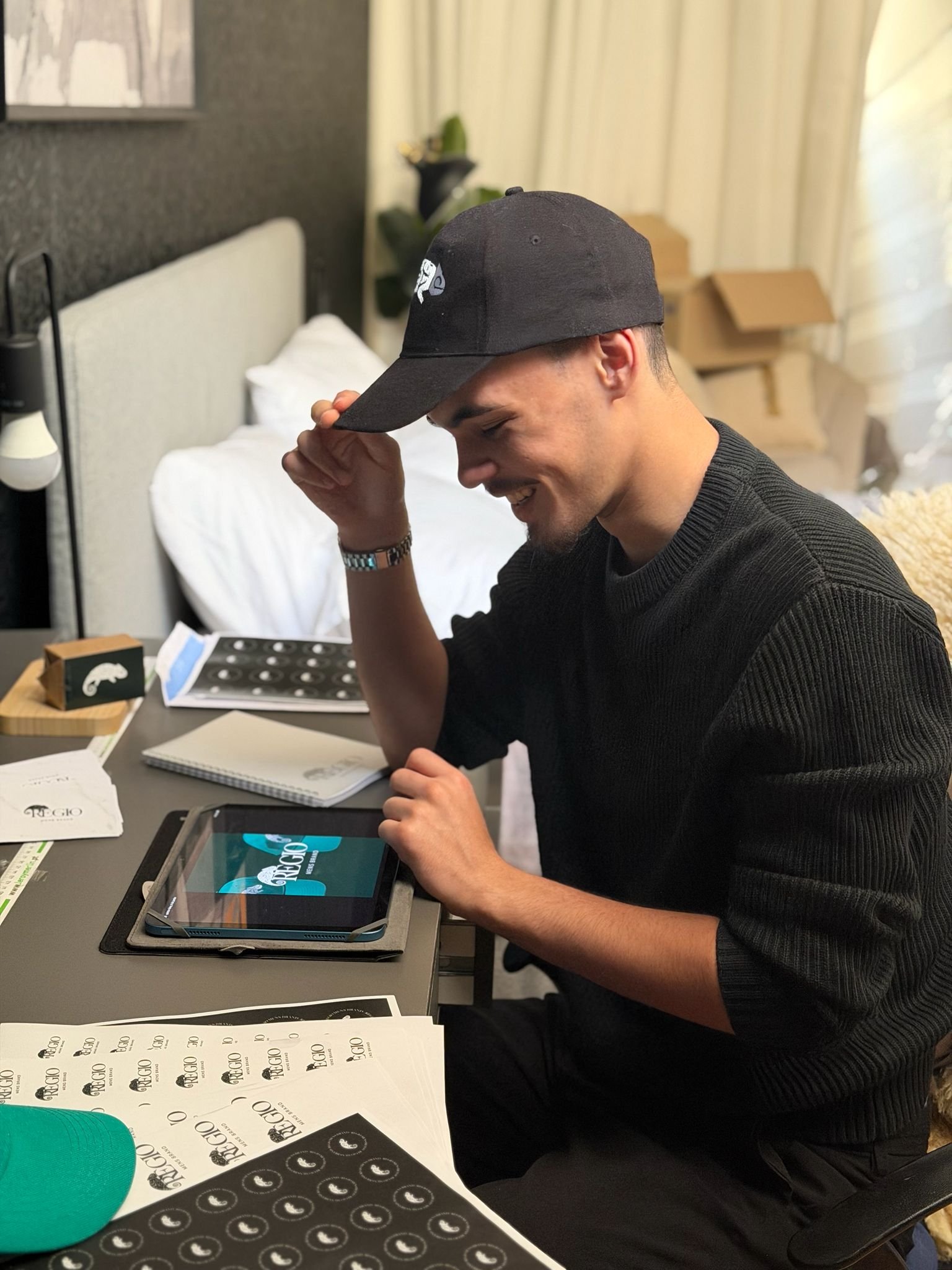 Homem sorridente usando boné preto, sentado à mesa com materiais impressos e um tablet que exibe o logo 'Prego'.