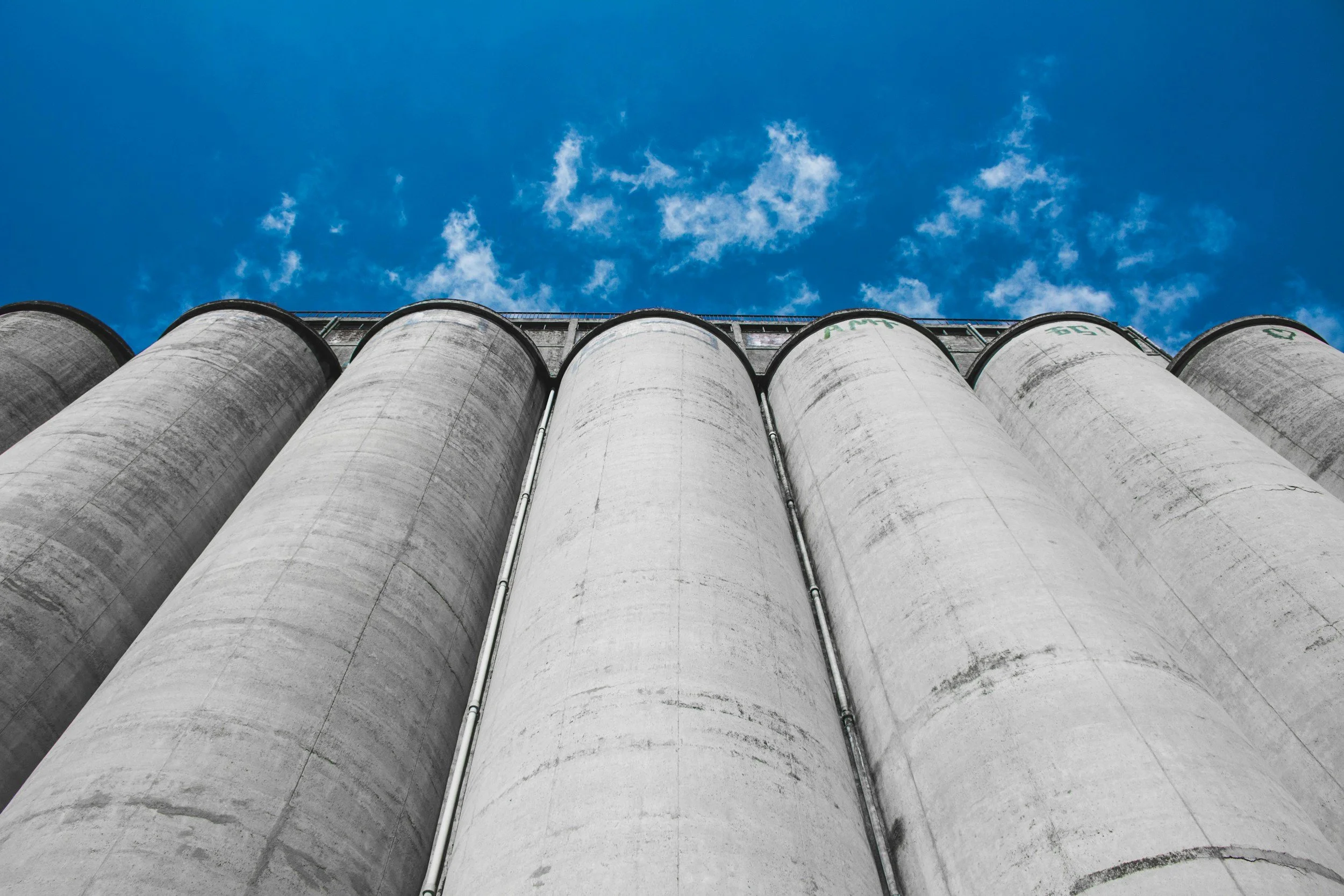 Six large industrial silos made of concrete against a bright blue sky with scattered clouds.