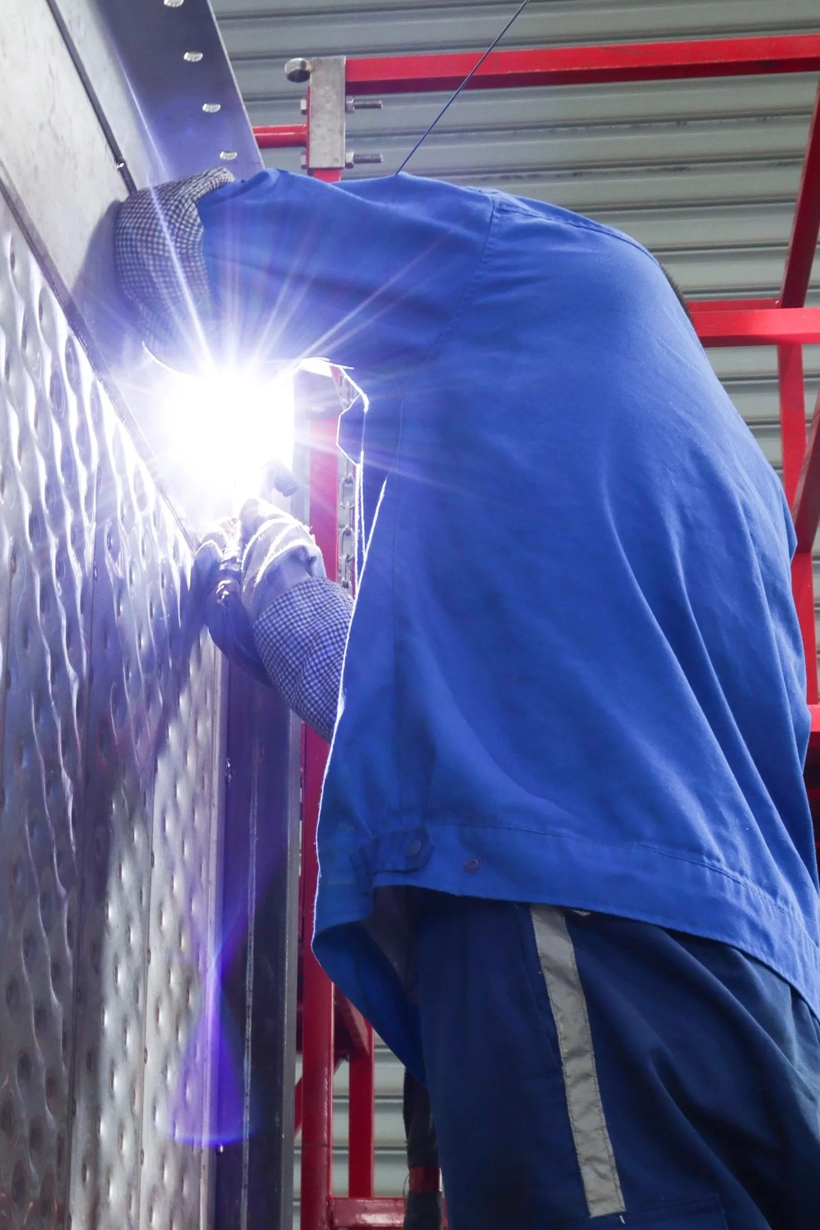 A person welding metal on a textured metal surface inside an industrial setting, with a red scaffolding and a corrugated metal ceiling visible.