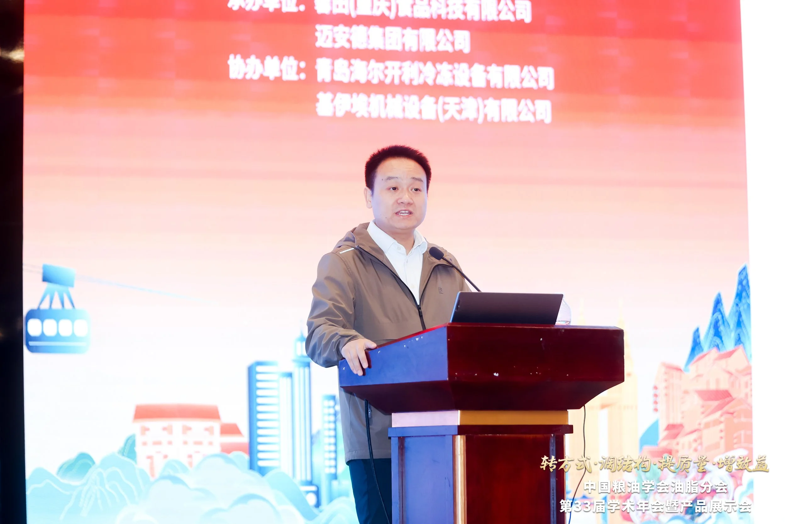 A man in a beige jacket speaking at a podium during a conference, with a large red screen behind him displaying Chinese text.