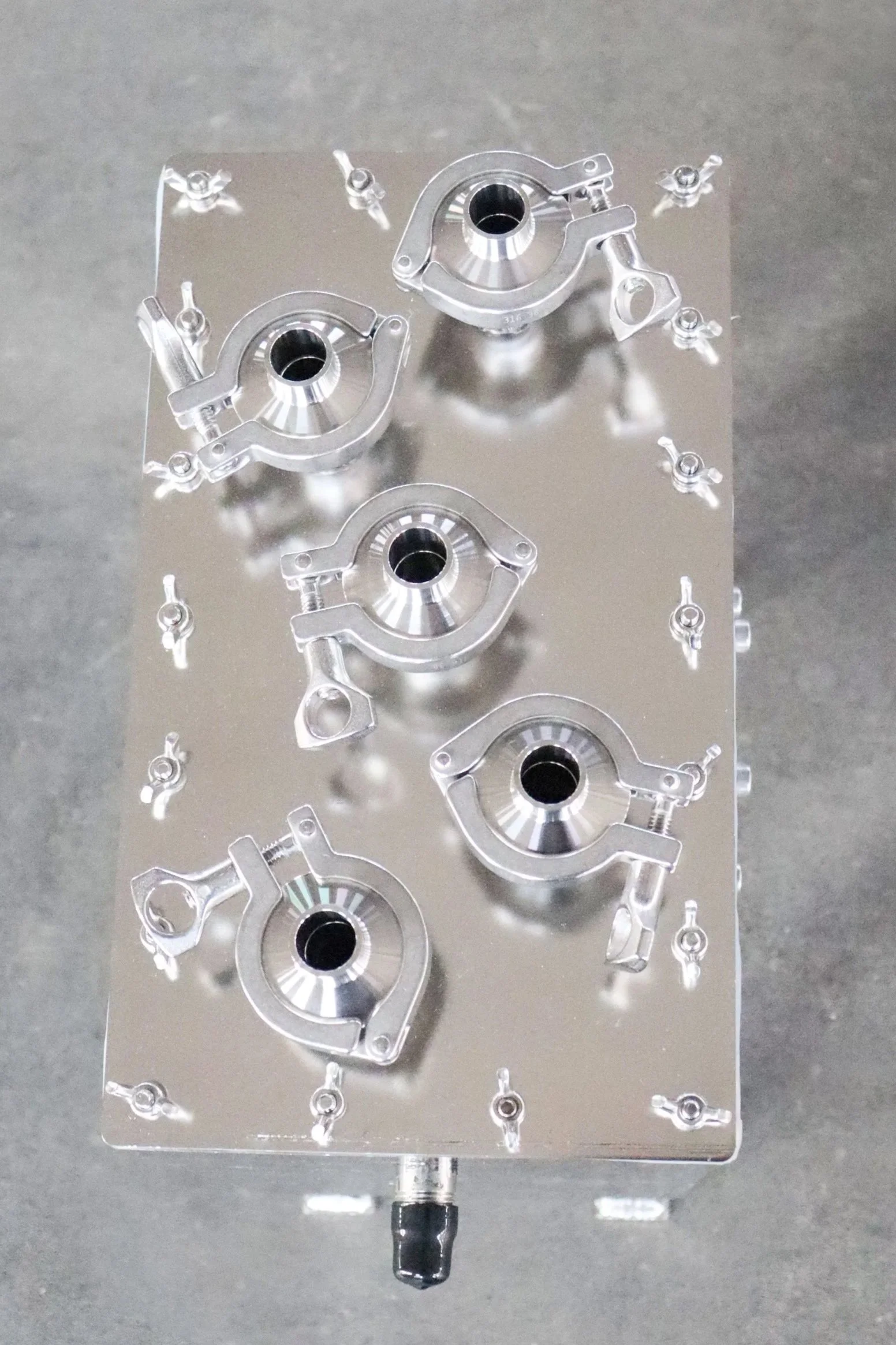 A metallic mechanical part with circular and angular components, mounted on a clear acrylic board with multiple small screws, placed on a gray surface.