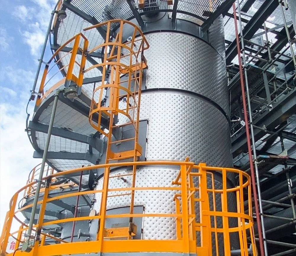 A large industrial building with metallic cylindrical structures, yellow safety railings, and a spiral staircase, set against a partly cloudy sky.