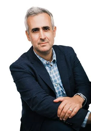 A man with gray hair wearing a dark blazer and a checkered shirt, standing with arms crossed against a white background.
