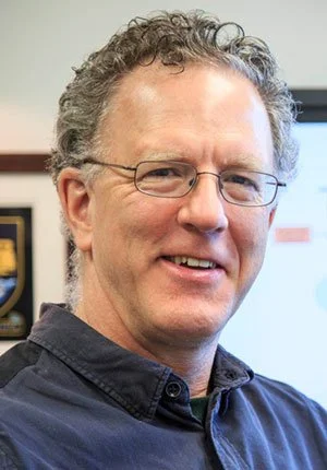 A man with curly gray hair and glasses, smiling, wearing a dark collared shirt.