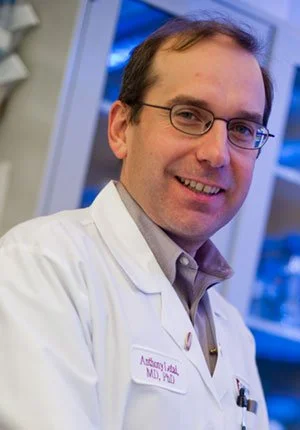 A man wearing glasses and a white medical coat with a name tag, smiling in a clinical setting.