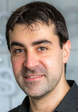 Close-up of a man with dark hair, brown eyes, and light stubble, smiling at the camera, in an indoor setting.