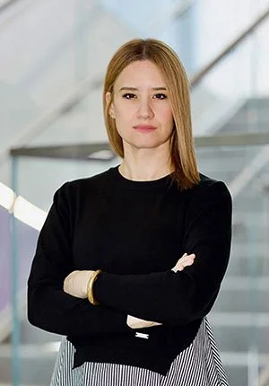 A woman with shoulder-length red hair wearing a black long-sleeve top standing with arms crossed in front of a modern building with glass windows.