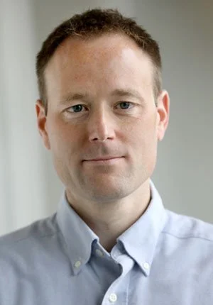 A portrait of a man with short brown hair, fair skin, wearing a light blue button-up shirt, looking directly at the camera with a neutral expression.