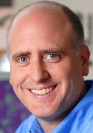 A man with blue eyes, short light brown hair, and wearing a blue shirt, smiling in an indoor setting.