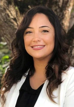 A woman with dark, wavy hair smiling outdoors near a tree.