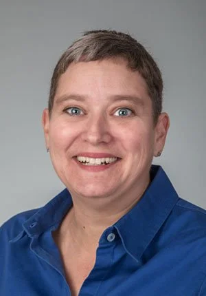Portrait of a smiling woman with short hair wearing a blue shirt against a neutral background.