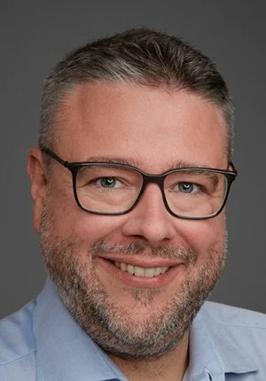 Close-up portrait of a smiling man with short gray hair, beard, glasses, wearing a light blue shirt, against a gray background.