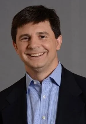 A man with dark hair, wearing a suit jacket and a light blue dress shirt, smiling at the camera against a plain gray background.