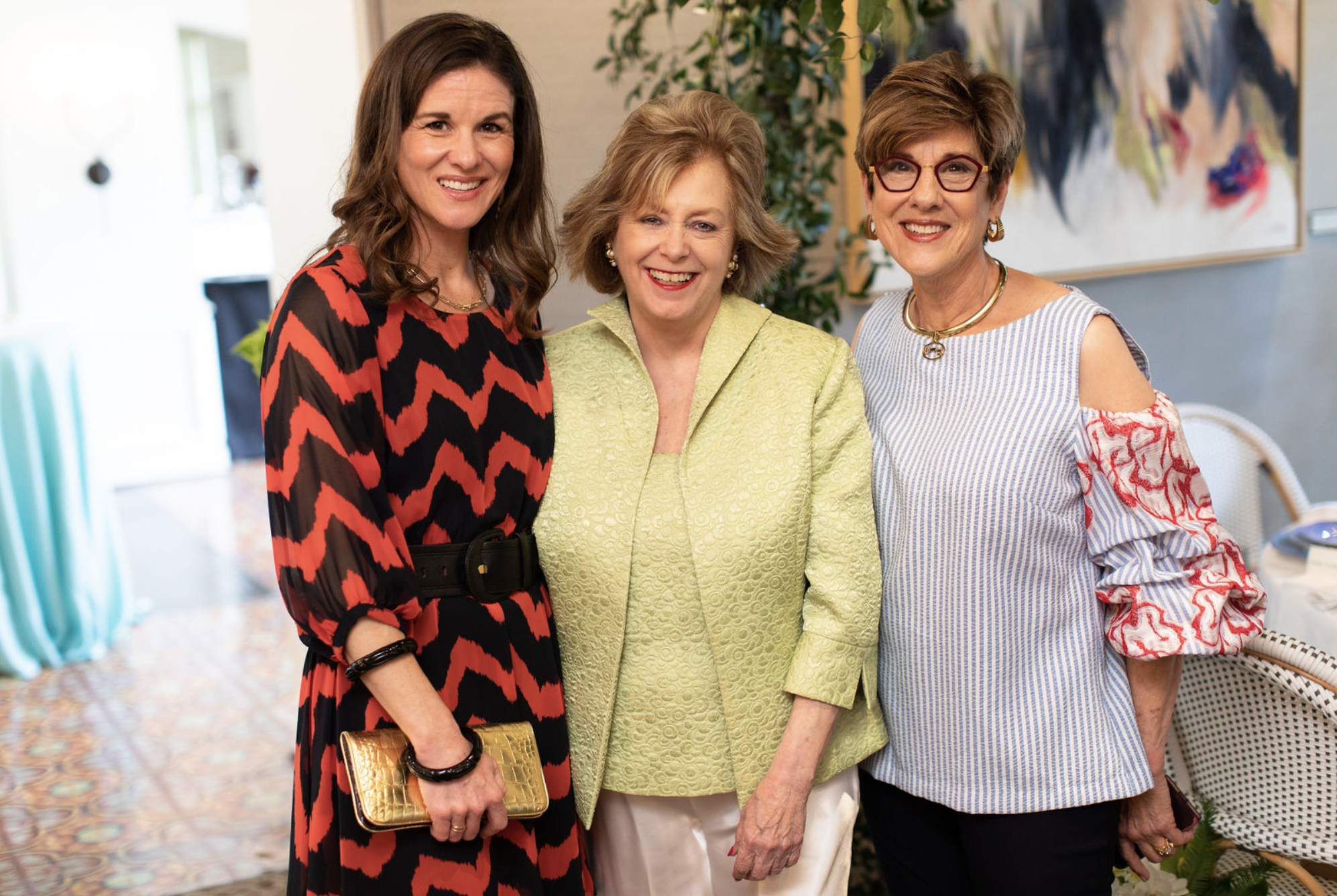 Three smiling women standing close together indoors, with art and plants in the background, dressed in colorful and patterned clothing.