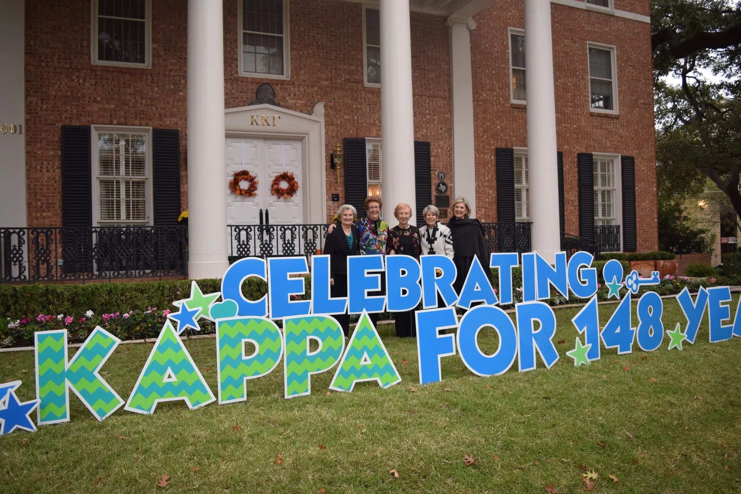 A group of five elderly women standing in front of a red brick house with white columns and black shutters, holding a colorful sign that reads 'Celebrating Family & Love for 148 years.' The house has two wreaths on the white door and a small porch wi