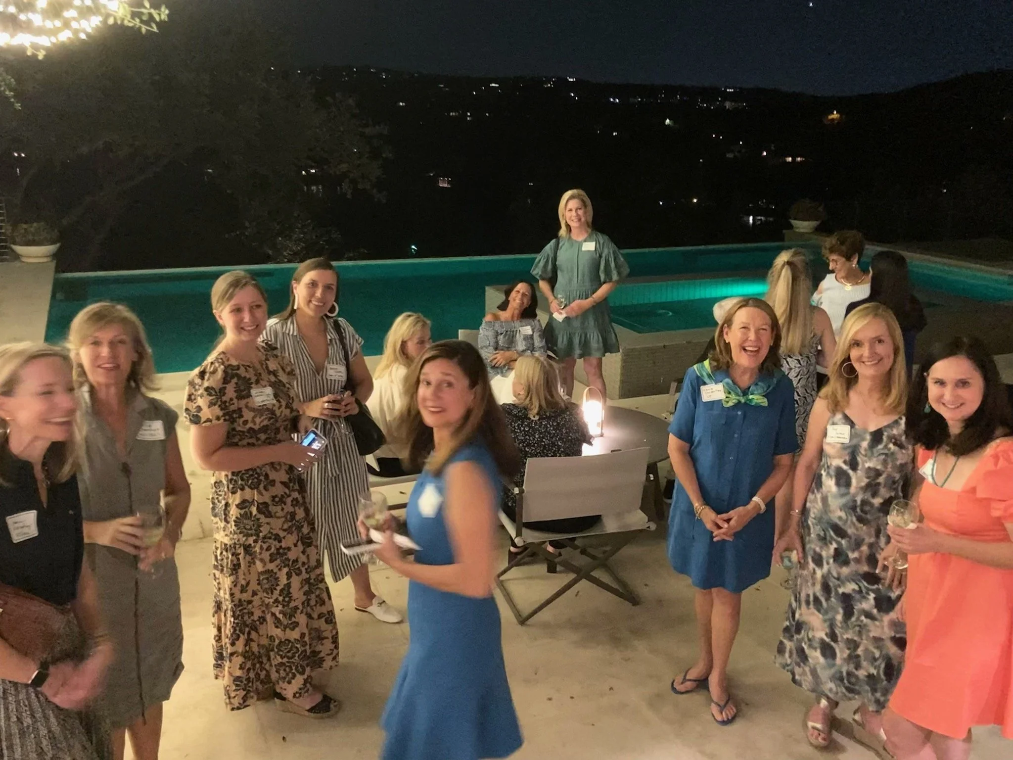 A group of women at an outdoor evening gathering around a pool, with some holding drinks and smiling.
