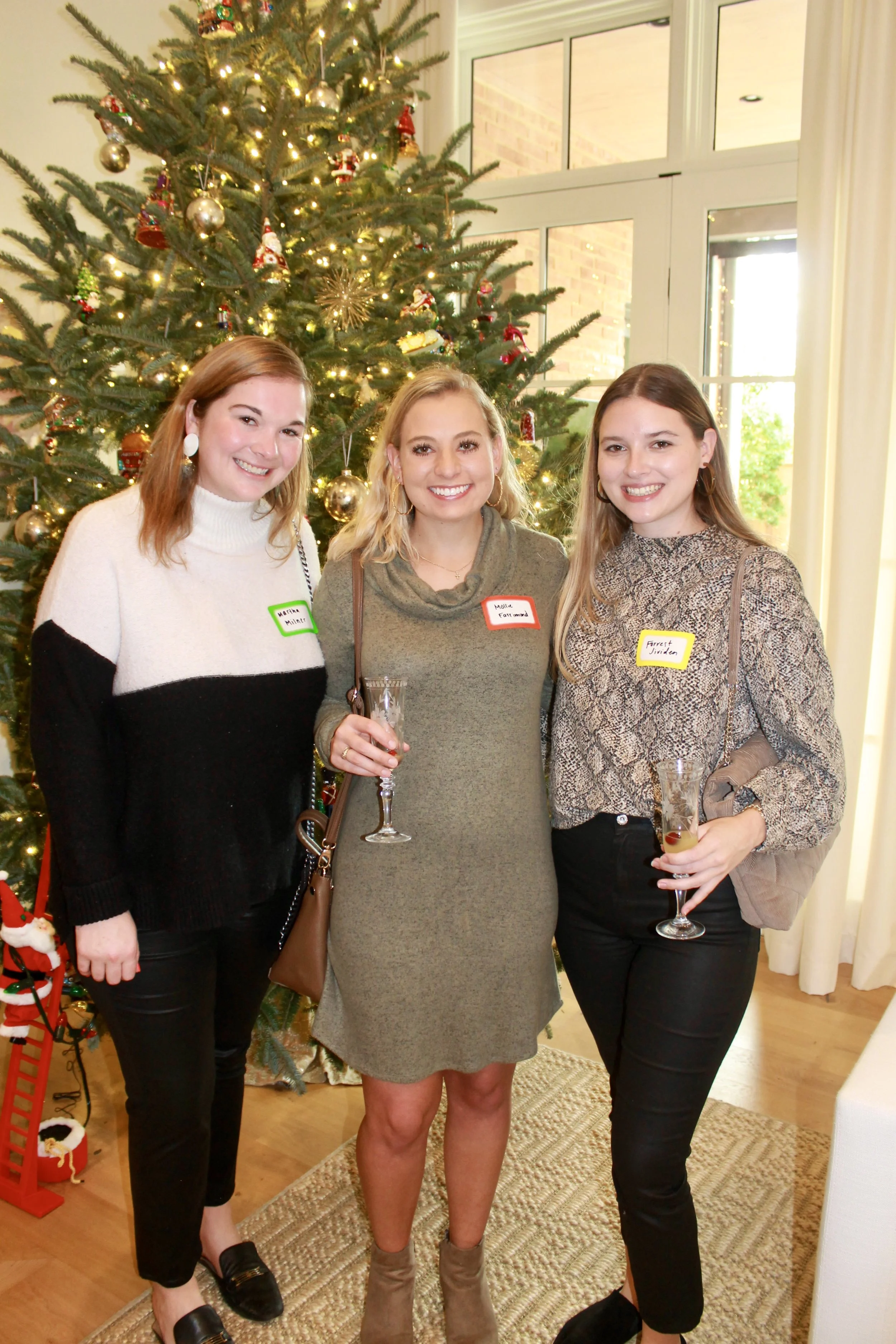 Three women standing in front of a decorated Christmas tree, smiling and holding drinks. They are wearing festive outfits and have name tags on their clothing.
