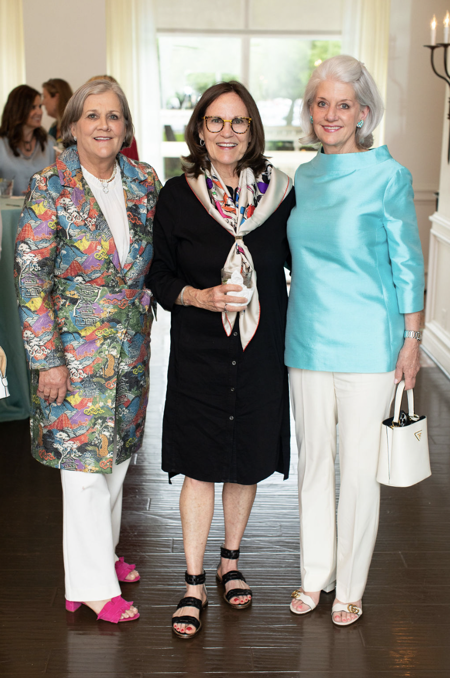 Three women dressed in colorful and elegant outfits, standing together indoors with a bright window and a candle sconce in the background.