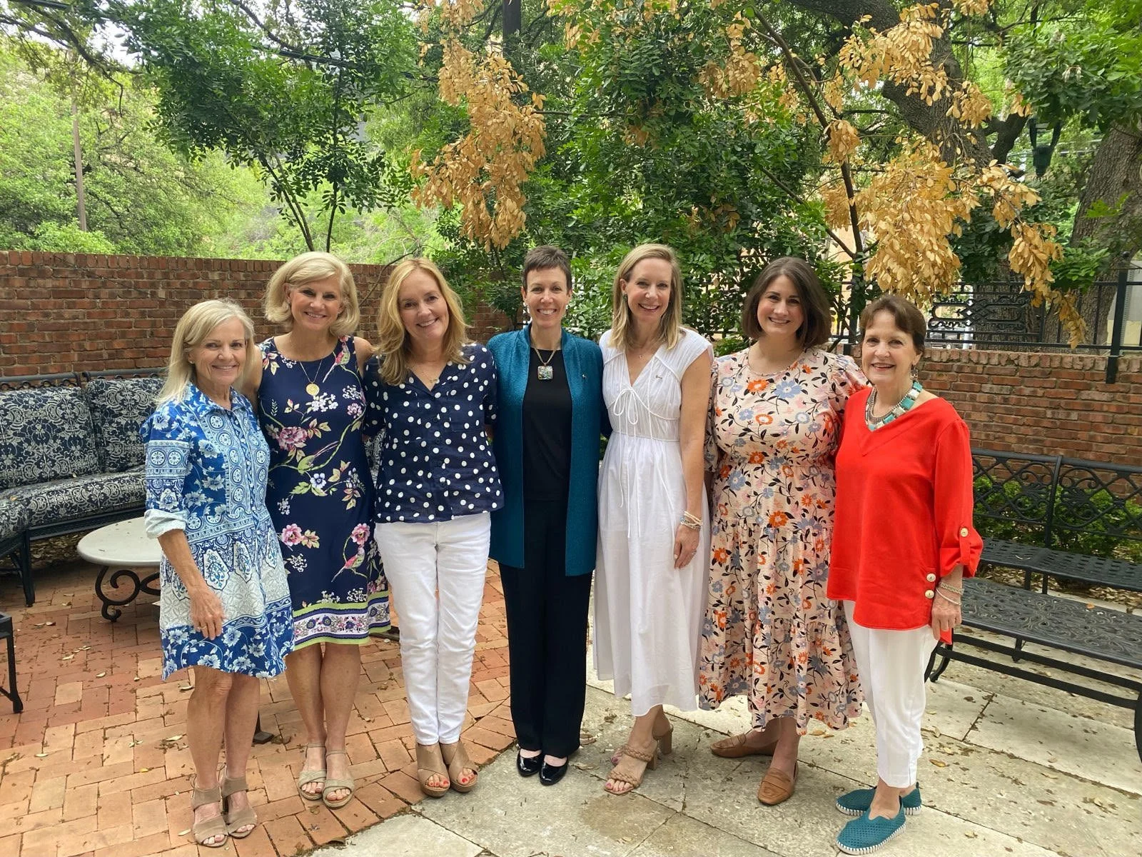 Seven women standing together outdoors on a brick patio, posing for a photo with green trees and a brick wall in the background. They are dressed in colorful summer dresses and smiling.