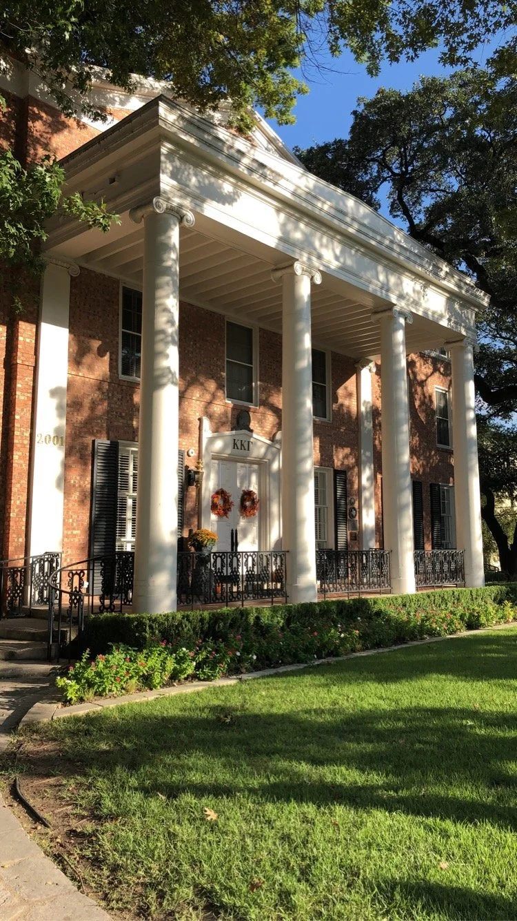 A large brick house with white columns supporting a porch roof. The house has black window shutters and a white door with the Greek letters KKI above it. There are autumn wreaths hanging on the door. The house is surrounded by green grass, bushes, and trees.