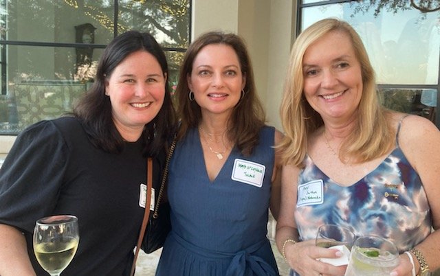 Three women smiling at an indoor event, each holding a drink, standing close together with name tags visible on their clothing.