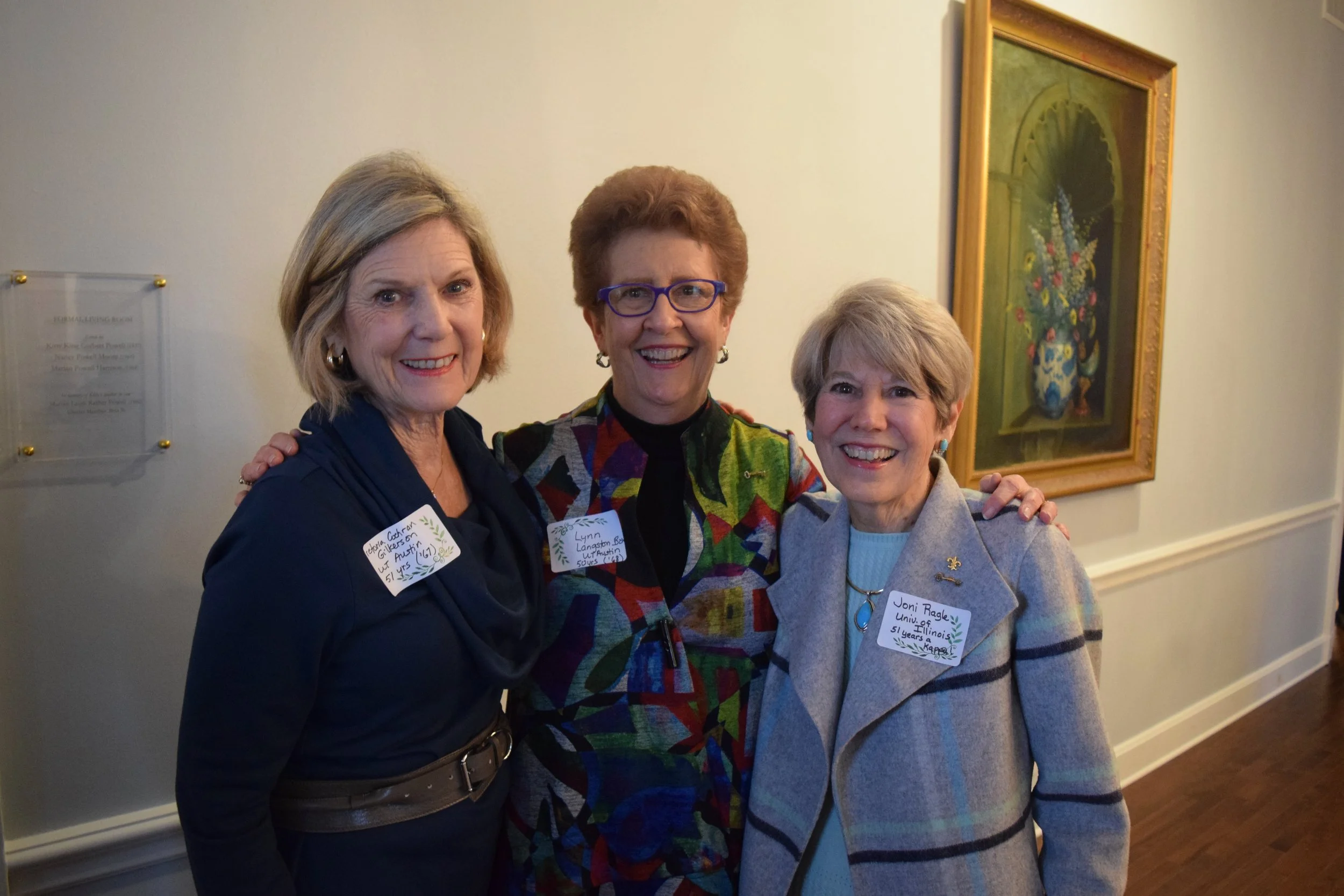 Three smiling women standing close together inside a room with framed art on the wall behind them. They are wearing name tags and casual clothing.