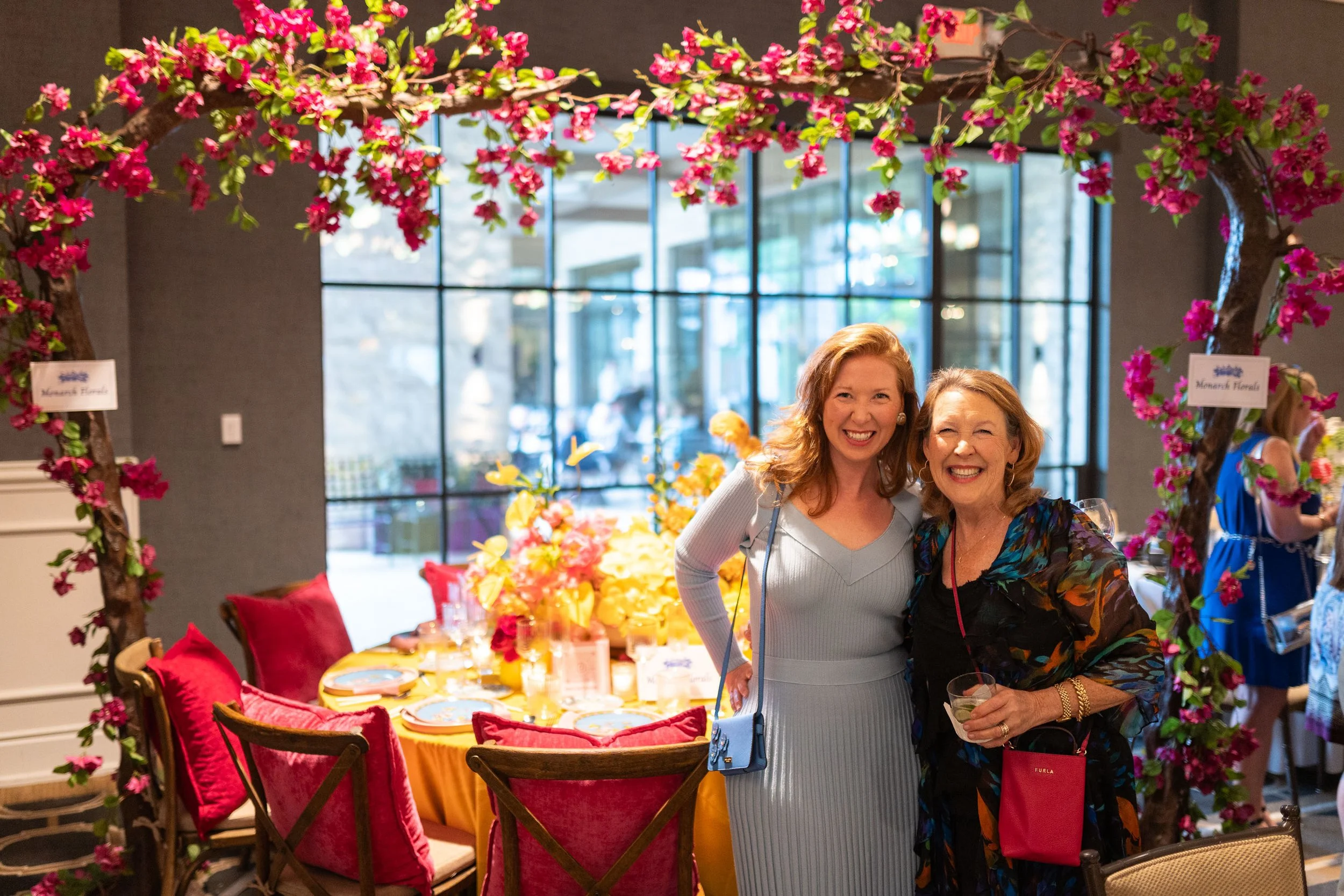 Two women smiling at a decorated indoor event, with pink floral arch framing the scene, a table set with colorful dishes and pillows, in front of large window panels.