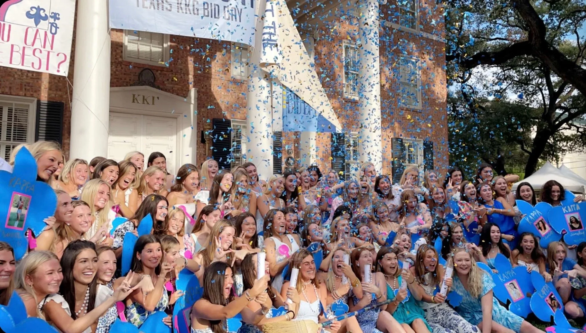 A large group of smiling young women gathered outdoors for a celebration, with blue and pink decorations and confetti falling in the air.