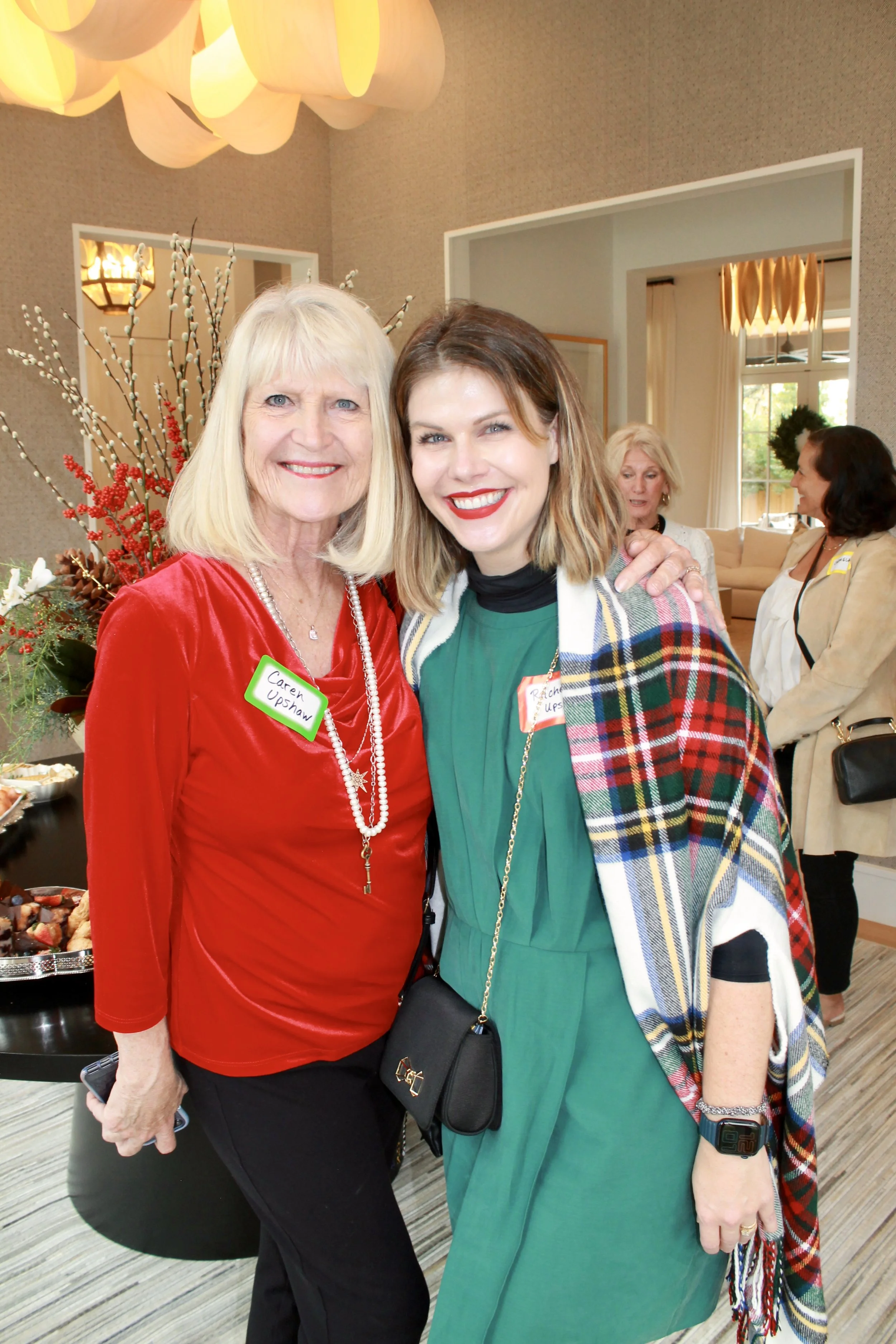 Two women smiling at a social gathering, one wearing a red top with a name tag that reads 'Carey Upshaw' and a necklace, the other wearing a green dress with a plaid shawl, and both have short hair. In the background, there are other women, a table w
