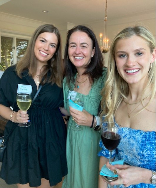 Three women smiling and holding drinks at an indoor social gathering.