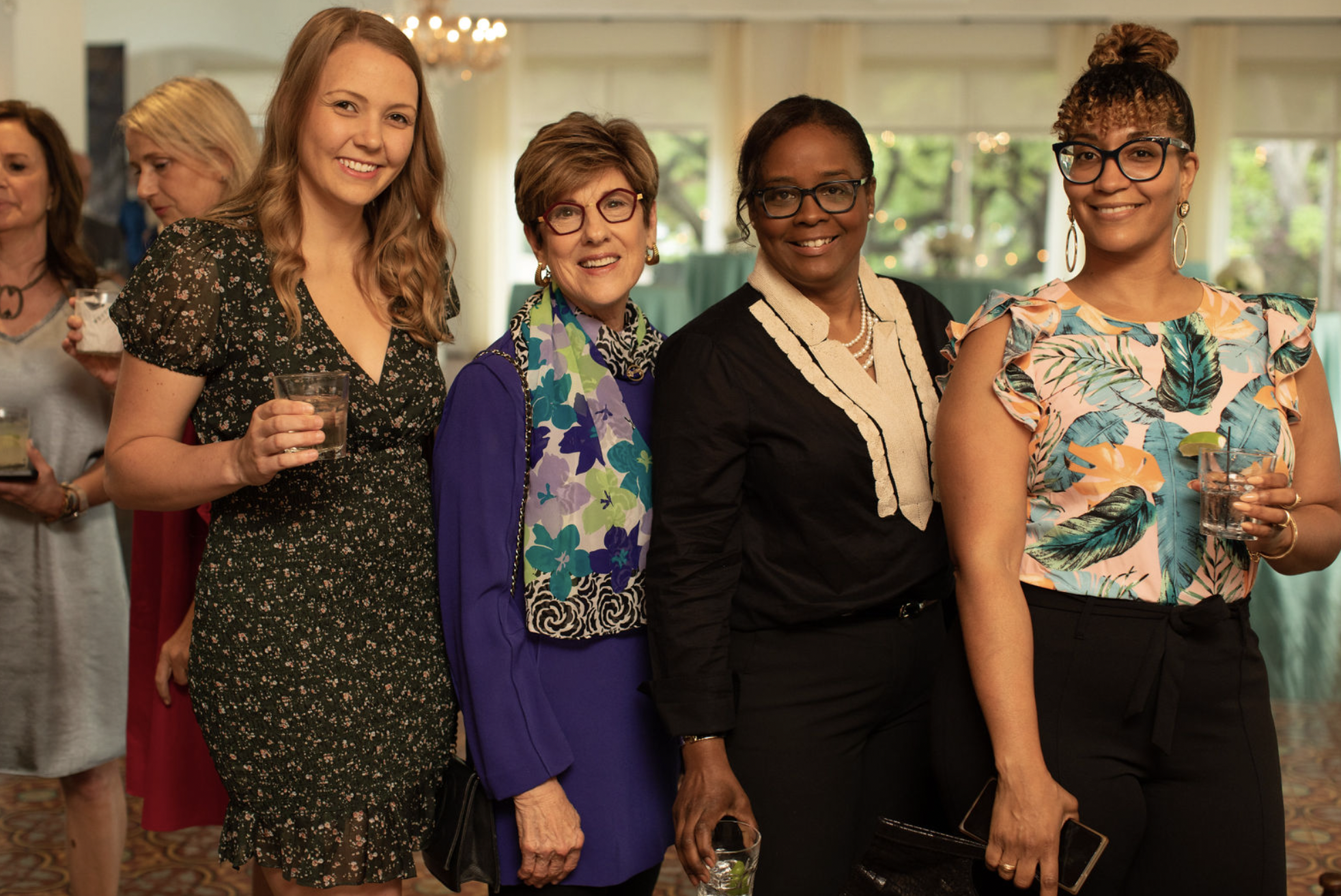 Four women at a social gathering, standing together and smiling at the camera, dressed in colorful and stylish outfits, with a bright, well-lit background.