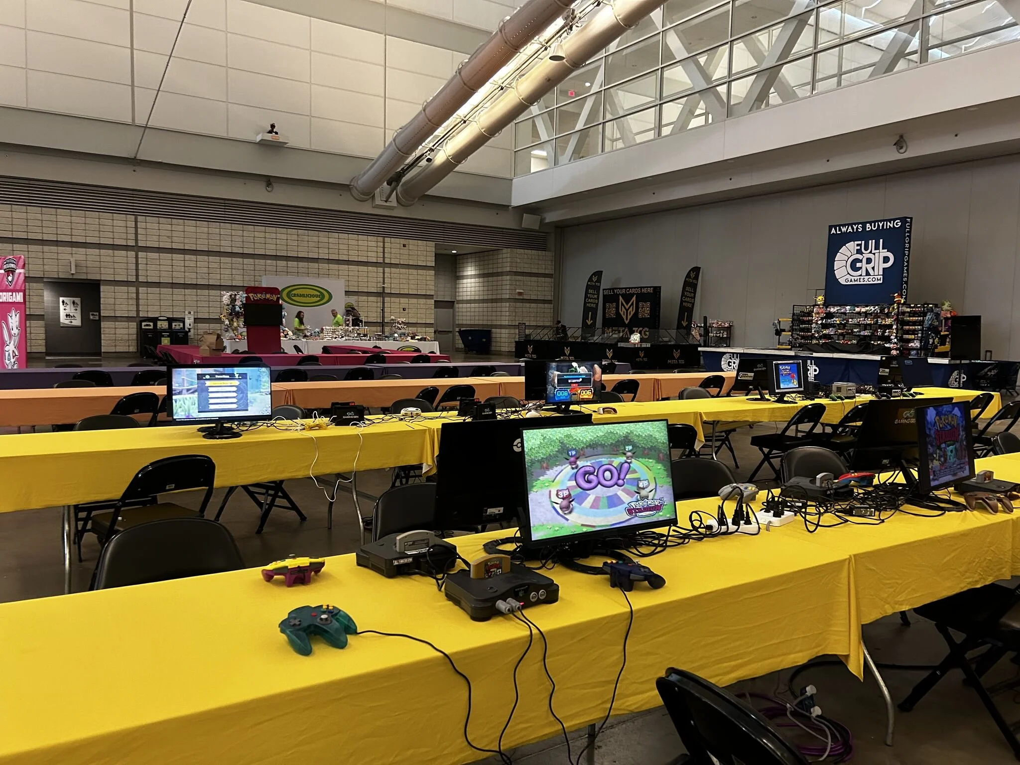 An indoor gaming event setup with yellow tables and black chairs, featuring multiple gaming monitors, controllers, and gaming consoles on the tables. In the background, there are vendor stands with signs, including one for Pokémon and another for Full Grip Games. The space has high ceilings with exposed ductwork and a second story with glass panels.