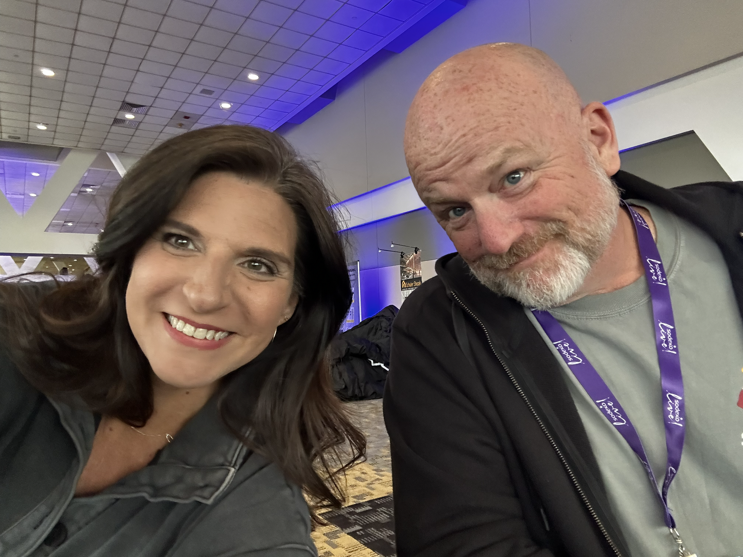 A woman with brown hair and a man with a bald head and gray beard taking a selfie at an indoor event with blue lighting and conference lanyards.