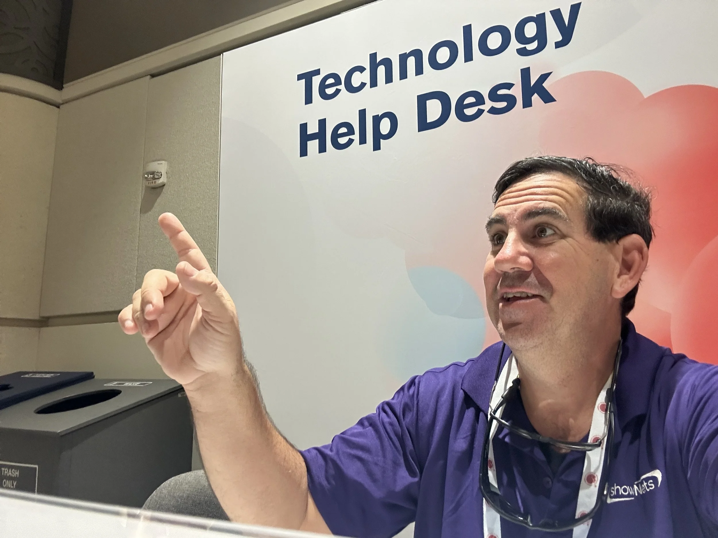 showNets Expo Floor Helpdesk. A man with dark hair and a beard, wearing a purple shirt with a shownets logo, pointing upward in front of a sign that reads 'Technology Help Desk.'