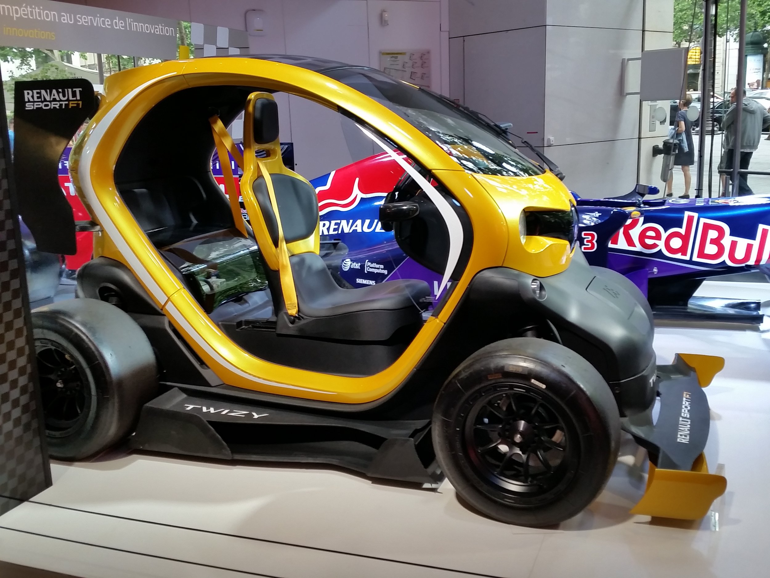 A Renault Twizy electric mini car with a yellow and black design displayed indoors, with racing sponsorship and branding including Red Bull and Renault Sport logos, and a transparent enclosure around it.