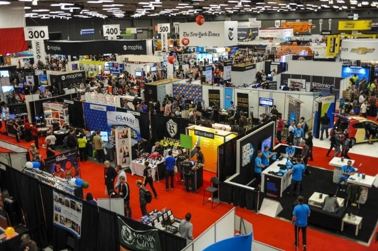 A crowded trade show or expo with various booths and displays, packed with attendees exploring different exhibitor setups and signage overhead.