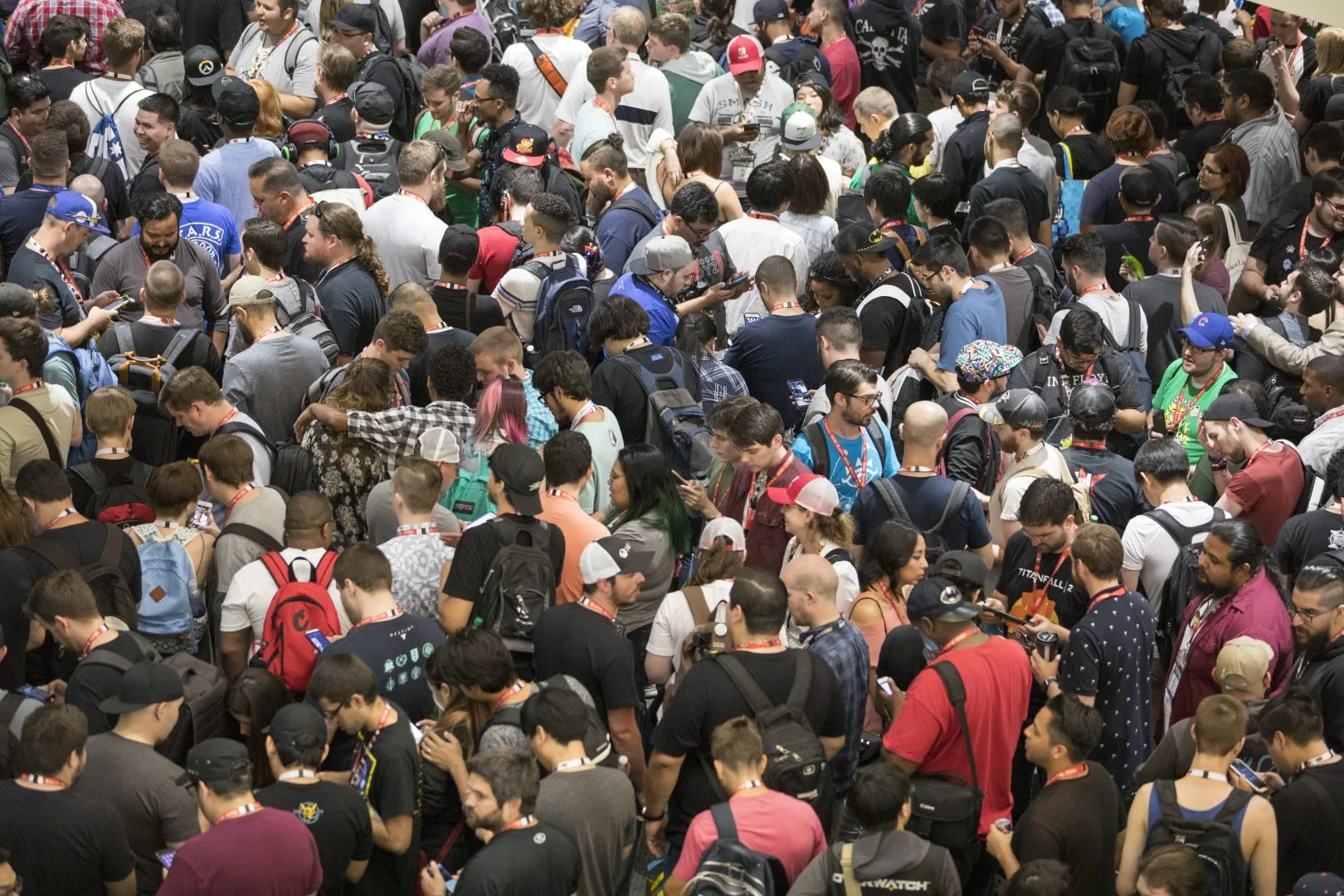 Crowd of people, mostly young adults, standing close together at a convention or event, many using smartphones and wearing backpacks.