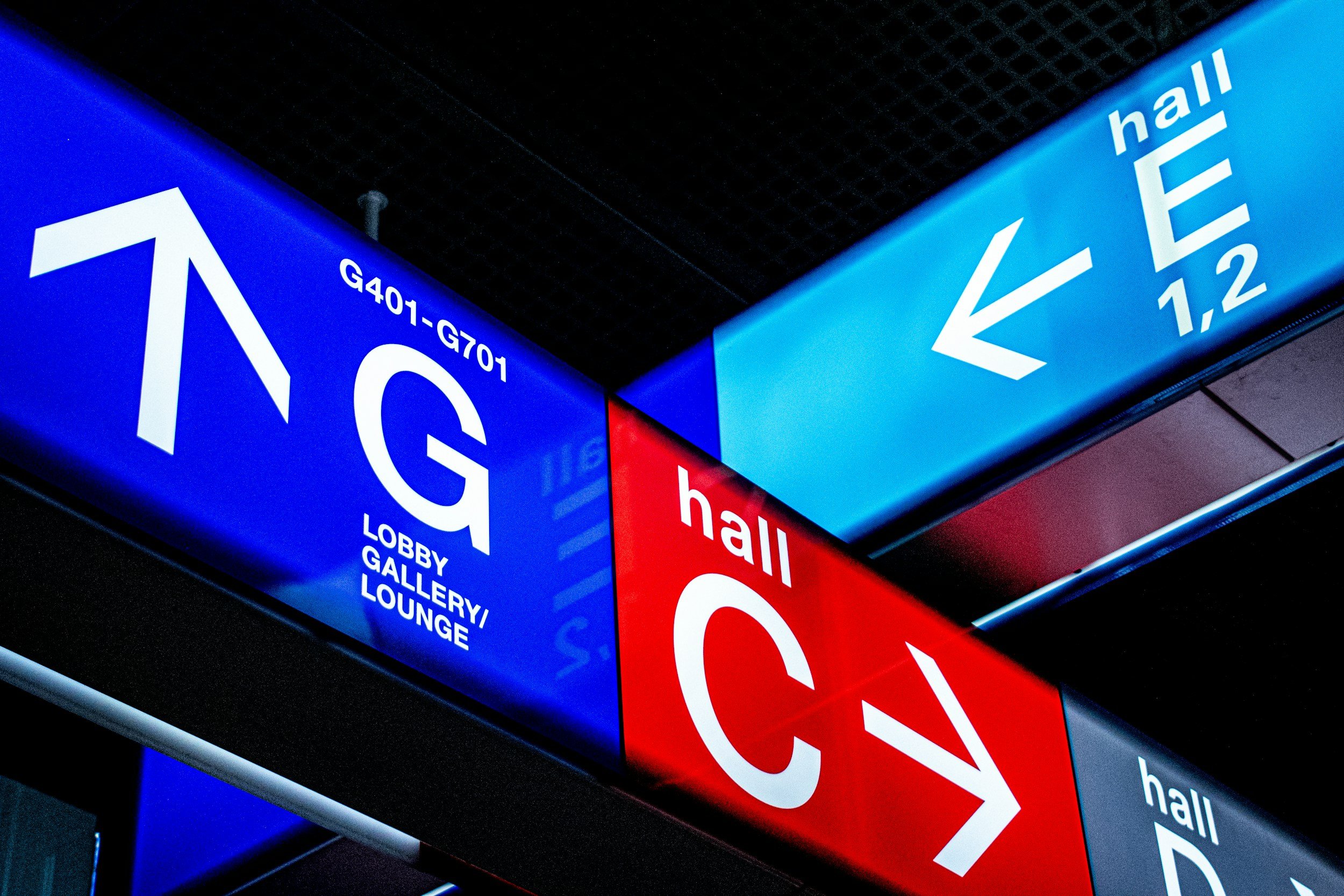 Blue and red illuminated signage with directional arrows and text indicating directions to hall, lobby, gallery, lounge, and gates G401-G701 in an airport.