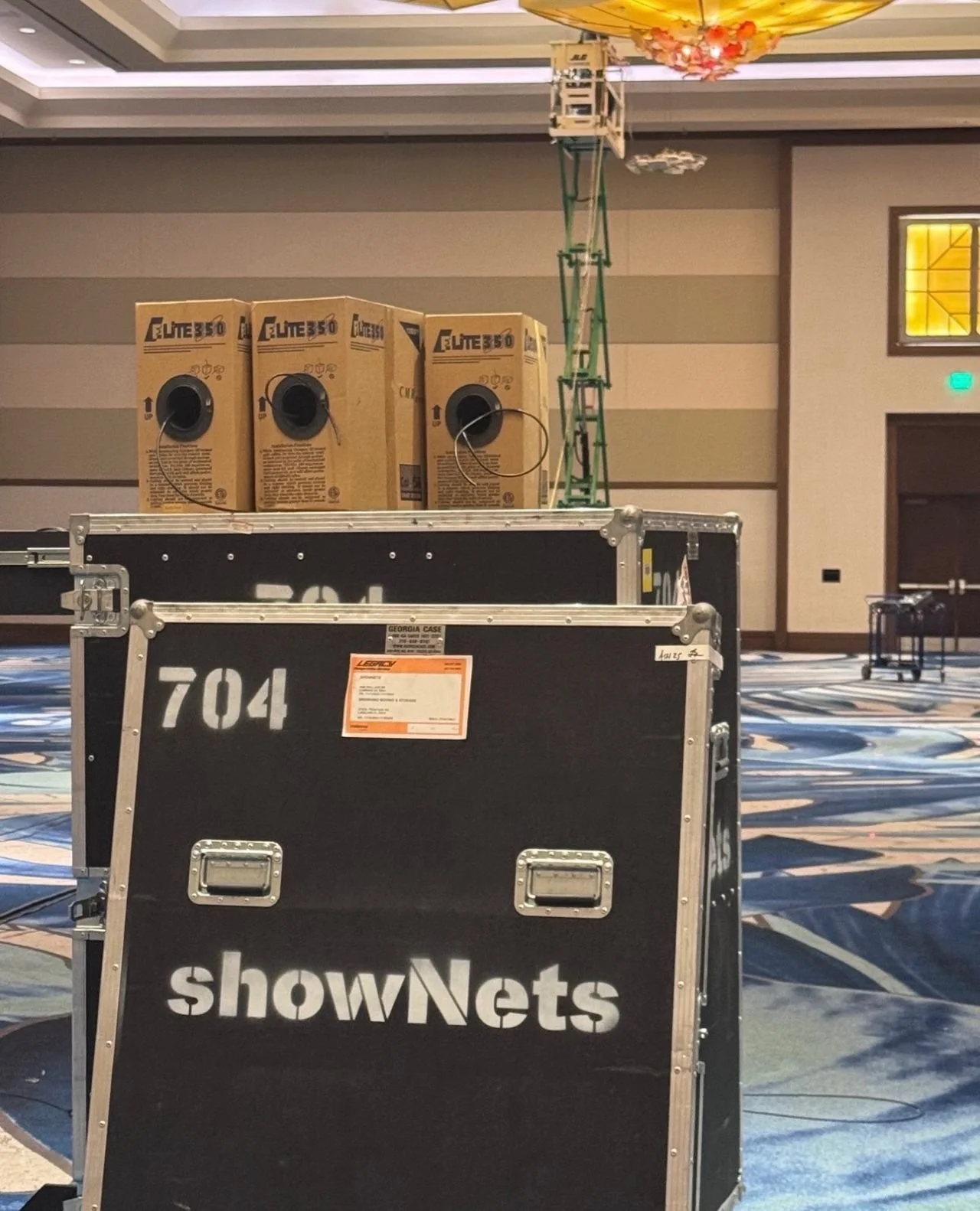 Boxes and a large black road case with showNets branding are set up in an empty conference room with decorative carpet and a ceiling chandelier. One box labeled Elite 350 is visible, and a green scaffolding or equipment stand is seen in the backgroun