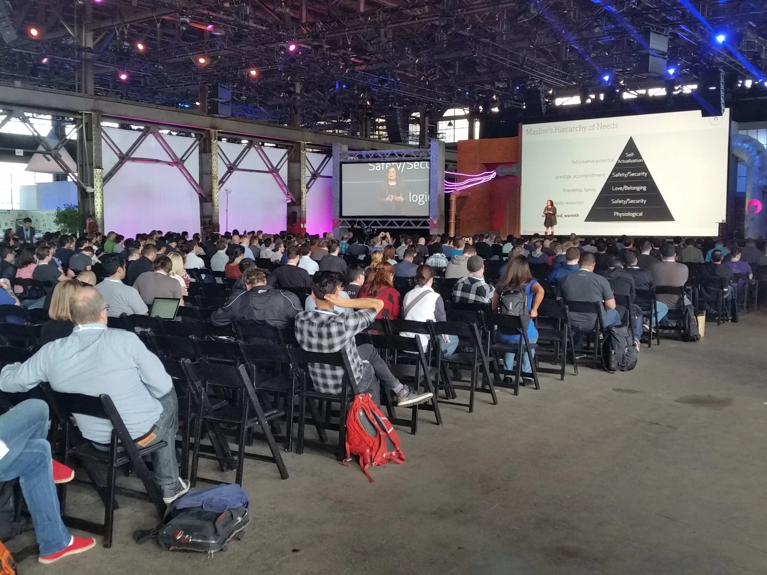 A large conference hall filled with seated attendees listening to a female speaker on stage with a presentation slide. The slide shows Maslow's Hierarchy of Needs in a pyramid format.
