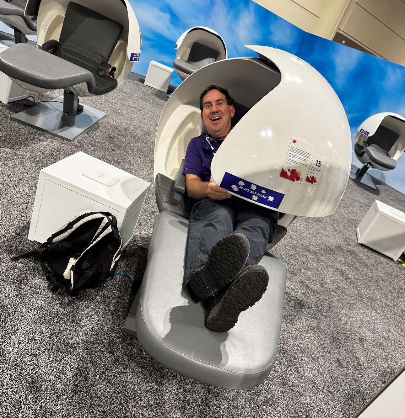 A man sitting inside a pod-style massage chair at an exhibit or store, holding a promotional pamphlet and smiling. The pod has a futuristic design with a rounded, enclosed shape. showNets Technician trying out the exhibits.
