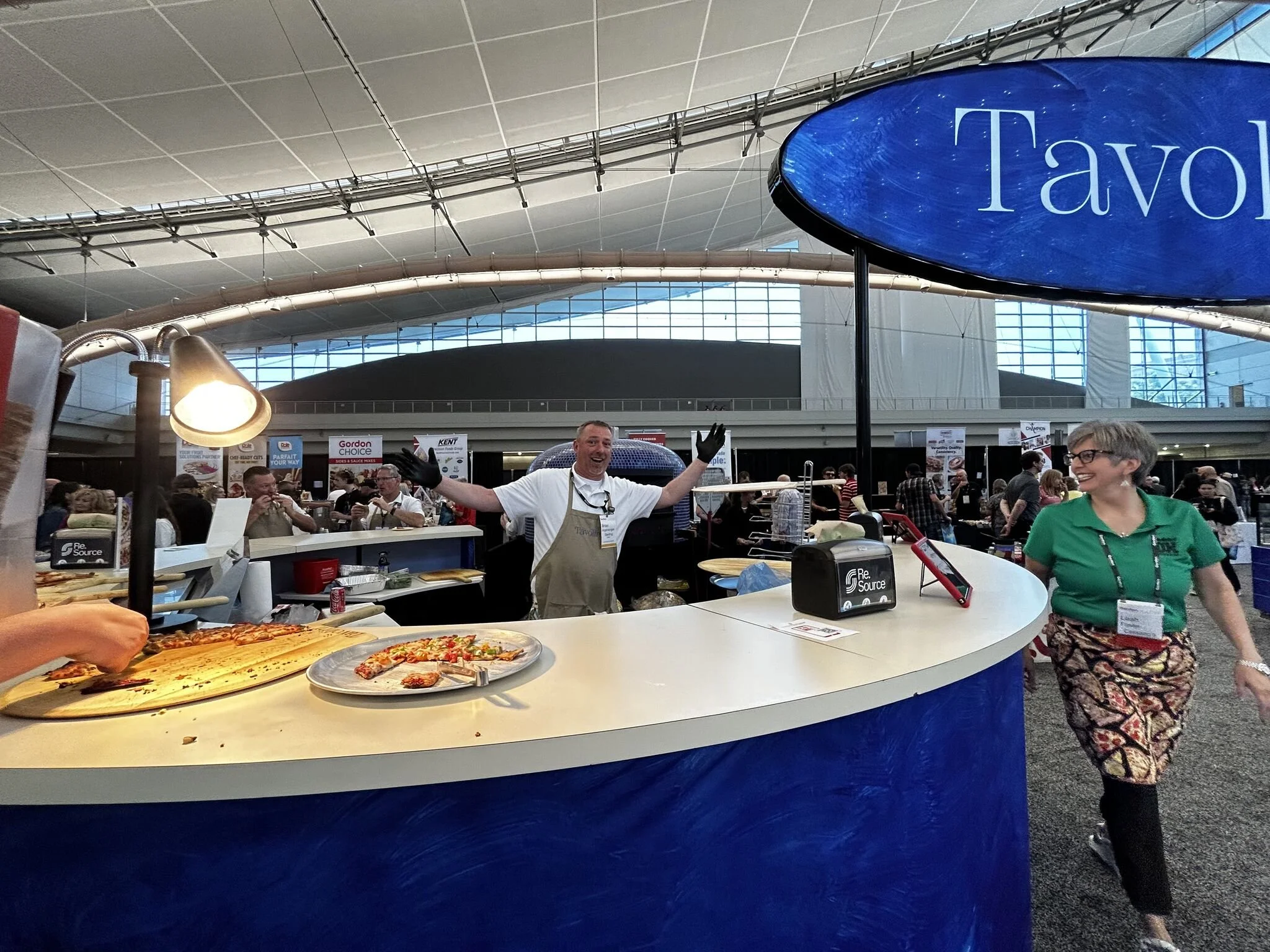 A busy food service booth at an airport with a large blue sign that reads 'Tavo'. There are several people behind the counter serving food and attendees in the background. Pizza is displayed on the counter in the foreground, and a man in a white shir