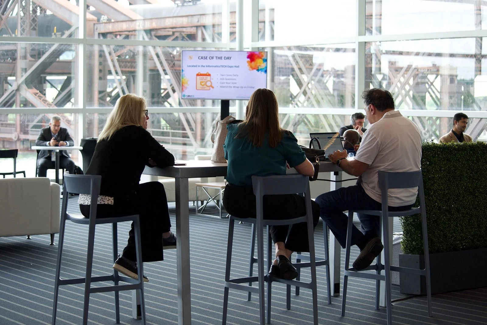 People working and using laptops at a high table in a modern office space with large glass windows and city views.